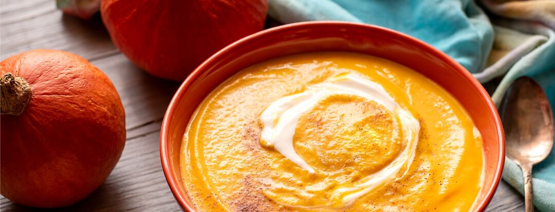 A bowl of soothing soup sits next to a pumpkin.