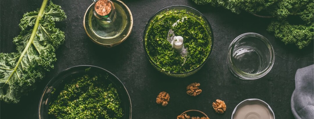 A blender full of pesto is surrounded by kale, garlic, walnuts and oil.