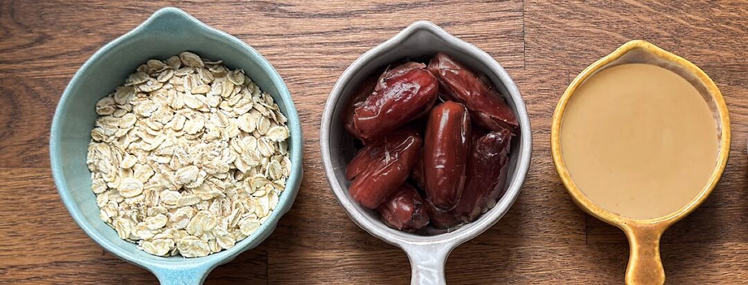 3 measuring cups for cookies sit on a wooden surface. They are full of ingredients; oats, dates, and Tahini.